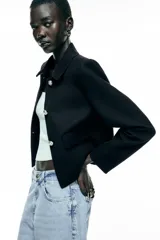 Chaqueta corta negra de tweed con cuello solapa, manga larga, bolsillos frontales con solapa y cierre frontal con botones plateados.