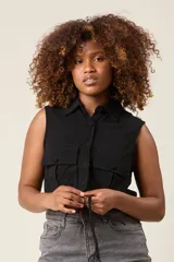 Chaleco cropped color crema, confeccionado en lino, con cuello camisero, cierre frontal con botones, dos bolsillos con solapa en el pecho y lazo ajustable en la cintura.