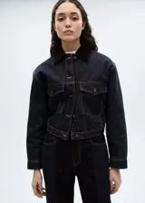 Chaqueta corta de denim azul oscuro con cuello camisero de pana marrón, cierre frontal con botones metálicos y dos bolsillos de parche con solapa abotonada en el pecho.
