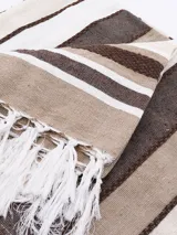 Pie de cama rectangular con flecos blancos en los extremos y diseño a rayas horizontales en tonos beige, marrón y blanco.