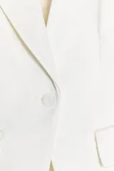 Blazer blanca de tejido satinado, con cuello solapa y escote pico, manga larga con hombreras, bolsillos con solapa en delantero y falso bolsillo en pecho, bajo con abertura en espalda y cierre frontal con botón.