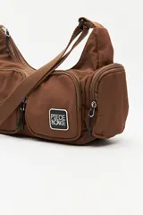 Cartera tipo shoulder color beige, de diseño utilitario y formato curvo. Confeccionada en material liviano y resistente, cuenta con correa regulable para llevar al hombro o cruzada. Presenta múltiples bolsillos con cierre en el frente y laterales, y un parche con el logo "Piece & Cake" en el frente.