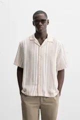 Camisa de manga corta con cuello bowling y corte relaxed fit. Diseño a rayas verticales en tonos beige y blanco. Cierre frontal con botones y bajo con aberturas laterales.