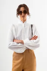 Camisa blanca de manga larga con cuello alto fruncido y puños ajustados. Presenta un corte holgado y tela con textura ligera.