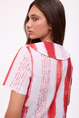 Blusa de manga corta con cuello camisero y botones, con estampado de rayas verticales rojas y texto manuscrito.