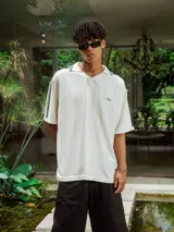 Chomba de tejido de punto cerrado color blanco, con cuello polo y mangas cortas. Presenta un bordado sutil en el frente y tres franjas negras en los hombros.