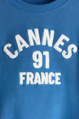 Buzo azul con cuello redondo, puños y bajo acanalados. Estampado frontal con la leyenda "Cannes 91 France" en color blanco.