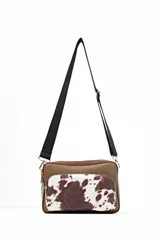 Bandolera rectangular con cuerpo principal de eco-piel texturizada color marrón topo y bolsillo frontal con estampado de vaca (marrón y blanco). Posee correa ajustable ancha de tela negra y herrajes dorados.