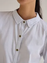 Camisa celeste de algodón con cuello clásico, cierre frontal con botones y mangas largas. Presenta volados en el frente y corte evasé.