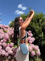 Bandolera pequeña tipo 'phone bag' de símil cuero color rosado pálido, con cierre de cremallera y correa ancha de tela con diseño de chevron en tonos claros.