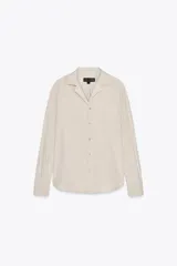 Camisa de manga larga color crudo, confeccionada en hilatura de algodón. Tiene cuello solapa, bolsillo de plastrón en el pecho, bajo redondeado y cierre frontal con botones.