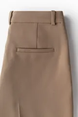 Pantalón de vestir color marrón claro, de tiro alto, con cierre y botón, bolsillos al bies y bolsillos insertados decorativos atrás. Piernas ajustadas con pliegue cosido adelante, raya planchada atrás y tajo corto en los bajos.