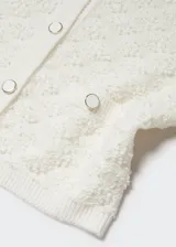 Cárdigan color crema con textura de punto, cuello redondo, cierre frontal con botones y bolsillos frontales decorativos. Corte cropped y mangas largas.