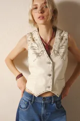 Chaleco de cuero de oveja color blanco hueso, de corte entallado, con cuello en V y cierre frontal de botones metálicos. Presenta bordados de tachas metálicas y piedras de colores en el frente, simulando motivos florales o de estrellas.