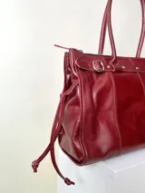 Bolso grande de cuero marrón con textura tipo cocodrilo, estructura rígida y asas para llevar al hombro. Tiene una tira con tachas y hebillas en cada extremo, también lleva lazos en los costados.