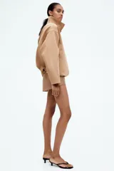 Campera color beige de sarga con cuello, cierre oculto adelante y bolsillos laterales con cierre. Modelo de corte holgado con pinzas en los codos para dar forma y cordón para ajustar elástico con topes en el bajo. Sin forrar.