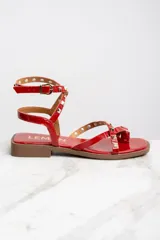 Sandalias rojas de charol con tiras finas, puntera cuadrada y cierre con hebilla al tobillo. Decoradas con tachas piramidales doradas.