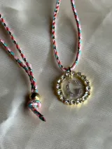 Collar con dije circular dorado con strass y letra 'A' en el centro, con cordón trenzado multicolor y cierre ajustable.