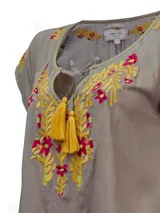 Blusa color taupe de mangas cortas con bordados florales en amarillo y fucsia. Tiene escote redondo con lazo y borlas amarillas.