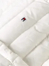 Campera blanca acolchada de nailon con relleno de plumas, cuello redondo, cierre con botones a presión, bolsillos laterales con botones a presión y logo de Tommy Hilfiger bordado en el pecho.