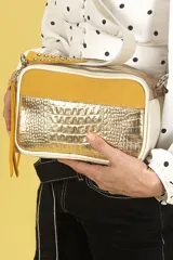 Bandolera rectangular con panel frontal de cuero croco metalizado dorado, laterales de descarne amarillo y bordes de napa offwhite. Tiene correa larga de cinta tejida offwhite con detalles en descarne y tachas metálicas, doble cierre superior y tiracierre de descarne con flecos largos.