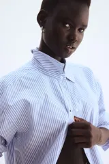 Camisa Oxford de algodón a rayas verticales celestes y blancas, con cuello clásico, botones frontales, canesú en la espalda, corte relajado, hombros caídos, mangas largas con puños sencillos y bajo redondeado con espalda más larga.