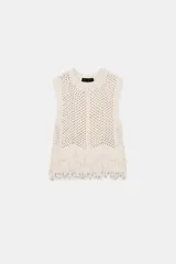 Chaleco de punto calado color crudo, de cuello redondo y sin mangas. El bajo está terminado con flecos.