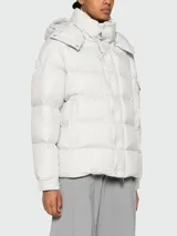 Campera puffer Moncler Maya color gris claro, rellena de plumas, con capucha desmontable, cierre frontal con botones a presión y bolsillos laterales con cierre.