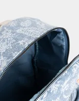 Mochila de denim azul con estampado paisley blanco, bolsillo frontal con cierre, correas regulables y manijas superiores de algodón con broche de cuerina. Acceso mediante cierre superior y puller de cuerina. Incluye tag de cuerina.