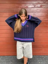 Sweater de punto trenzado color azul marino, con cuello en V y ribetes a rayas rojas y azules. Tiene un logo bordado en el pecho y aberturas laterales en el ruedo.