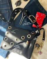 Cartera bandolera de cuero negro con ojalillos metálicos plateados, correa regulable con hebilla, bolsillo interior para labial y gancho para llaves. Diseño uruguayo.