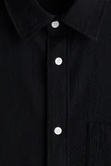 Camisa negra de sirsaca de algodón con cuello inglés, tapeta clásica, bolsillo superior de parche y canesú con pliegue de caja en la espalda. Mangas largas con tapeta de manga y botón, y cierre ajustable de botón en los puños. Bajo redondeado y corte holgado.