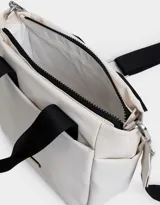 Cartera tipo tote color blanco hueso, con doble asa corta negra y correa larga negra regulable y desmontable. Tiene cierre superior, bolsillos laterales y tag de goma con el logo de la marca.