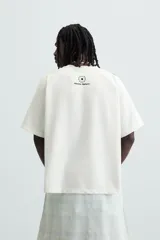 Camiseta blanca de corte holgado con mangas cortas negras. Presenta un logo azul en el pecho izquierdo, un escudo rojo con un caballo en el pecho derecho y la inscripción "Victory Games" en negro en el centro.