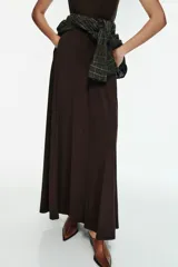 Vestido largo color marrón oscuro, con cuello alto y corte al cuerpo en la parte superior, y falda acampanada con pliegues. Presenta tirantes finos que dejan los hombros al descubierto.