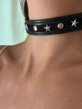 Gargantilla tipo choker de cuero sintético negro, con apliques de estrellas y remaches plateados. Se cierra con cadena y mosquetón para ajustar el largo.