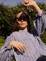 Camisa verde con rayas verticales blancas y bordados de flores. Tiene cuello mao, mangas largas abullonadas con puños elásticos y cierre frontal con botones de nácar.