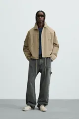 Cazadora color arena de corte relaxed fit, confeccionada en tejido de algodón. Cuenta con cuello solapa, manga larga con bolsillo multifunción, bolsillos frontales de plastrón, bolsillo interior, acabados con elástico y bajo ajustable con cordón. Cierre frontal con cremallera oculta por solapa con botones a presión.