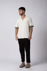 Camisa tejida color crudo, de manga corta, con cuello bowling y botones frontales. Presenta un tejido con textura de trenzas verticales y rib negro en los puños.