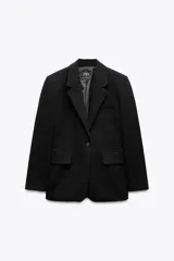 Blazer negra de corte clásico, con cuello de solapa y escote en pico. Tiene mangas largas con hombreras, bolsillos con solapa en el delantero y un bolsillo de vivo en el pecho. Está forrada y se cierra con un botón frontal.