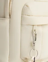 Mochila color crema de tela acolchada con pespuntes verticales. Tiene un bolsillo frontal con cierre, doble cierre superior, manija superior, correas regulables y bolsillos laterales. Incluye un charm colgante de corazón.