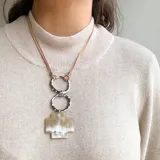 Collar con cordón de cuero marrón, dos aros metálicos color plata y dije de cruz color crema con detalles en blanco.