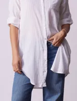 Camisola larga de algodón color blanco, marca Dash. Tiene cuello con solapa, abertura central con botones, bolsillo aplicado en el pecho, mangas regulables con presilla y botón, y bolsillos laterales en la costura.