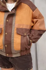 Chaqueta estilo bomber de gamuza con bloques de color en tonos tierra: naranja quemado, marrón oscuro y marrón claro. Presenta detalles en cuero liso en los hombros y bolsillos frontales con tapa y broche dorado. El puño y la cintura son ajustados con elástico.