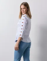 Camisa blanca de algodón con cuello clásico, solapa y escote en V. Tiene mangas tres cuartos con detalle de aberturas cruzadas. Presenta dos bolsillos delanteros aplicados y laterales en tejido modal.