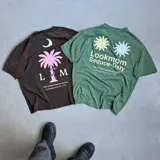 Camiseta marrón de corte oversize, con estampa en serigrafía bicolor (blanco y rosa) de una palmera, una luna y el logo de la marca.