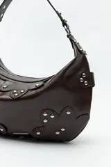 Cartera marrón de cuero sintético con forma de media luna, asa corta y apliques de tachas metálicas plateadas.