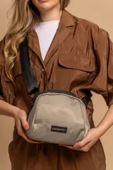 Bandolera pequeña de tela tafeta color beige claro, con correa ancha de nylon negro ajustable. Posee cierre superior negro.