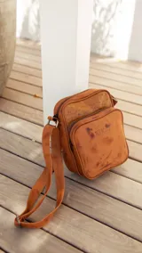 Bandolera de cuero color terracota con correa ajustable, cierre superior y bolsillo externo con cierre. Incluye bolsillo interno y logo de la marca en el frente.