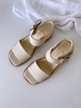 Sandalias de cuero color crudo, con tira ancha sobre los dedos, pulsera al tobillo con hebilla metálica y taco de madera de 6 cm de altura.